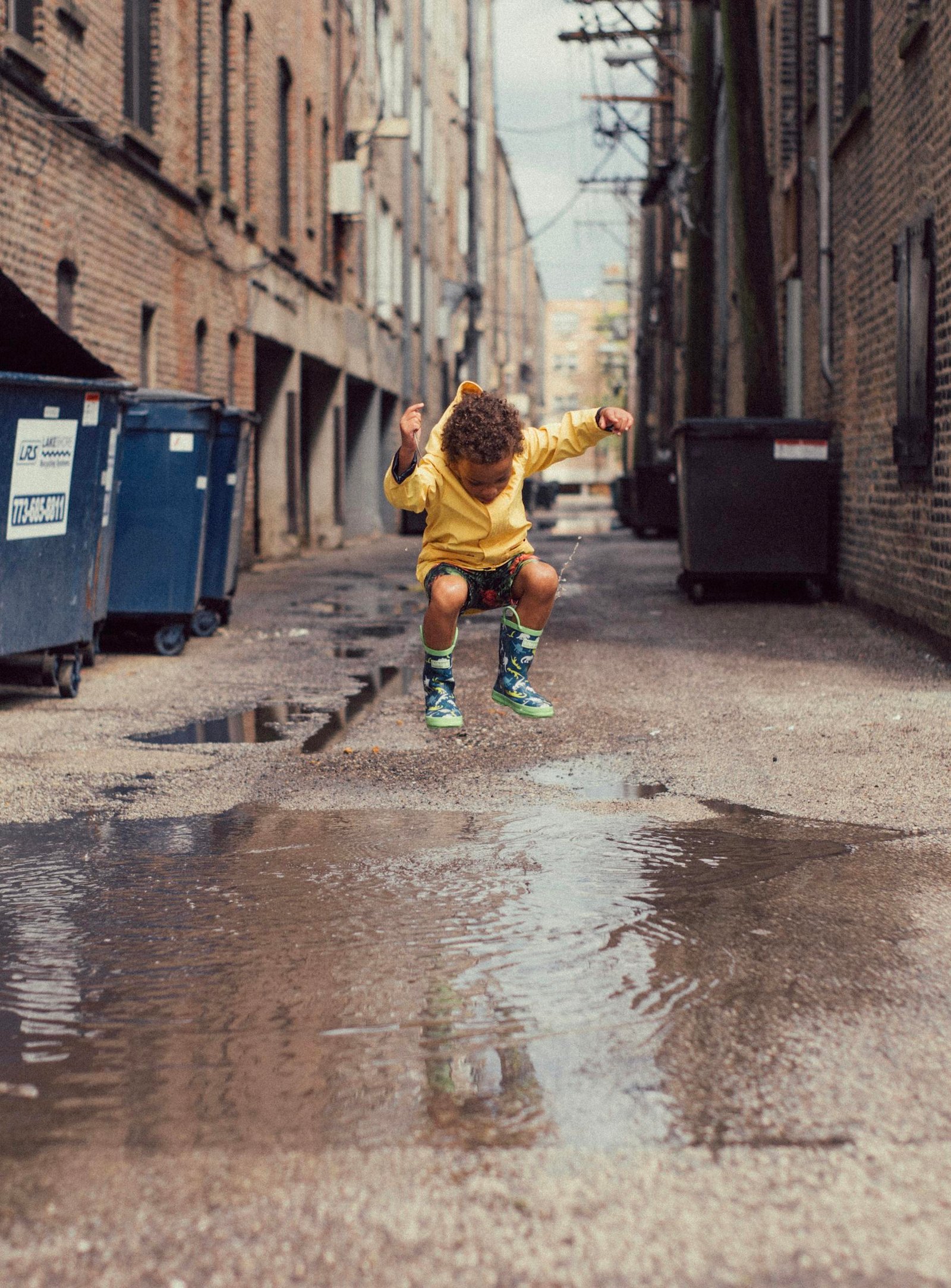 A young boy in a yellow outfit jumps over a puddle in an urban alley. Vibrant and playful moment.